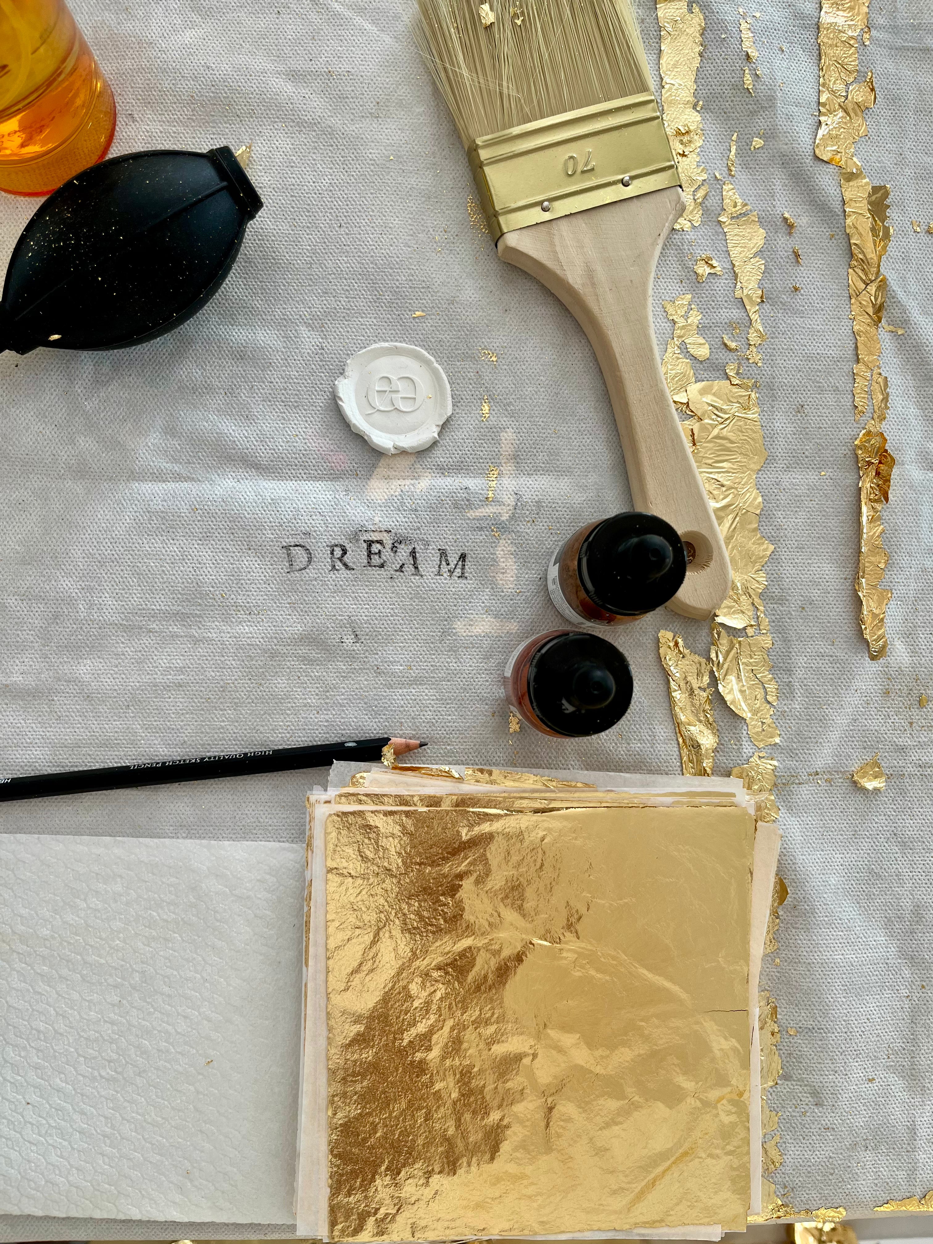Arbeitsmaterialien, wie Pinsel, Blattgoldimitatblätter, Bleistift usw. Im Mittelpunkt ist das Wort "Dream" auf die Unterlage gestempelt.