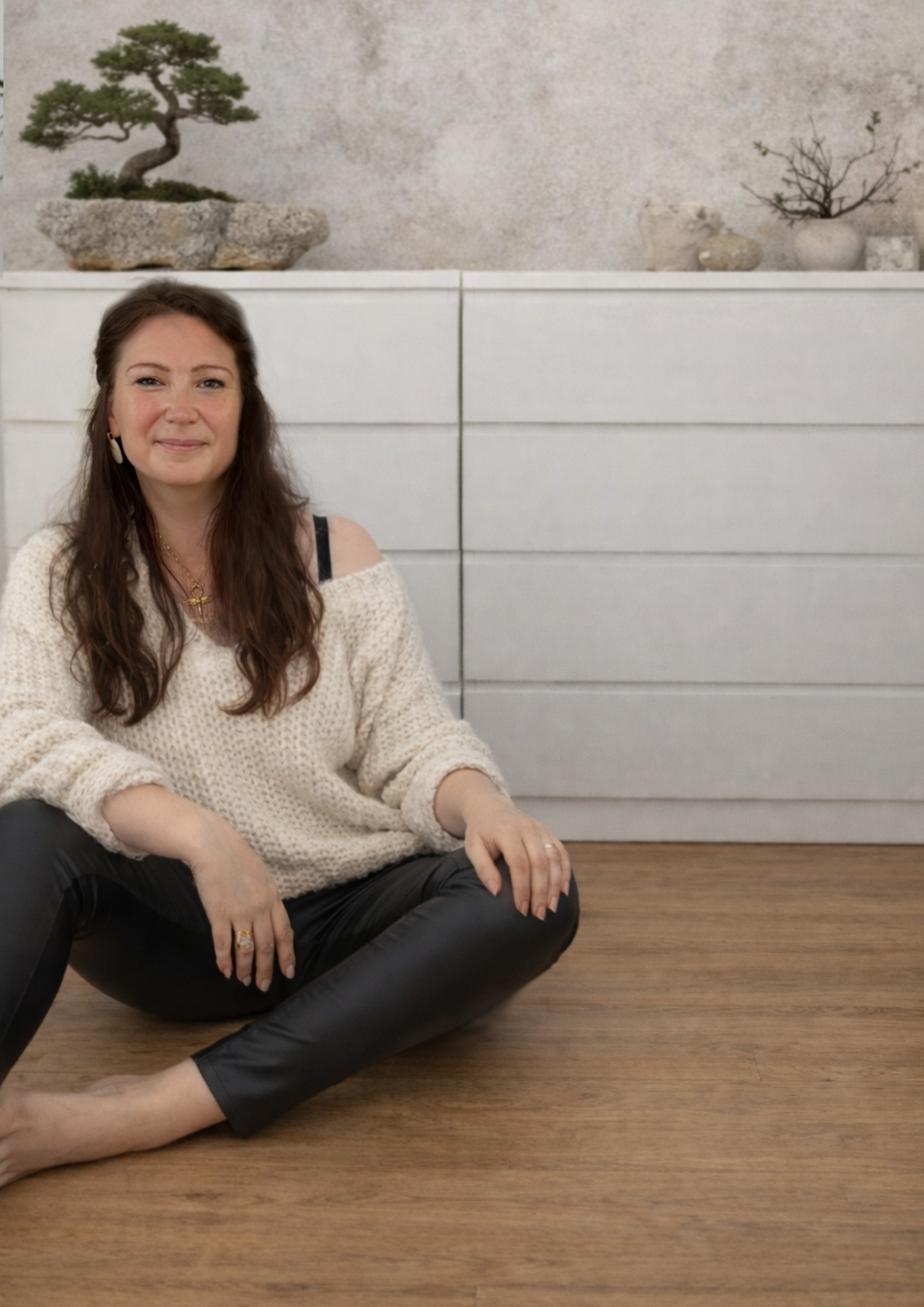 Nadine Elsner sanft lächelnd in beigem, weichen Pullover und Kunstlederhose. Braune lange Haare. Sitzend vor weißer Kommode auf braunem Parkett.