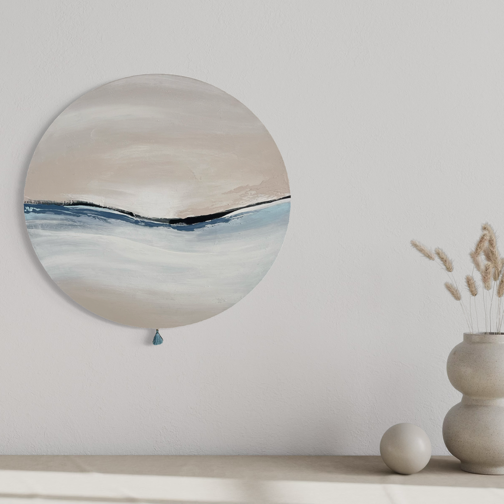 Das Kunstwerk "calm breath". Moderne Wabi Sabi Kunst mit daoistischer Seele von Nadine Elsner. 60cm im Durchmesser. Weiche nude, weiß und kühle blau Töne mit weichem Kunstlederstreifen. Sanfter Abschluss mit einer kleinen blauen Quaste. Das Bild gibt einem hellen Raum über einer beigen Kommode und Deko eine warme und klärende Atmosphäre.