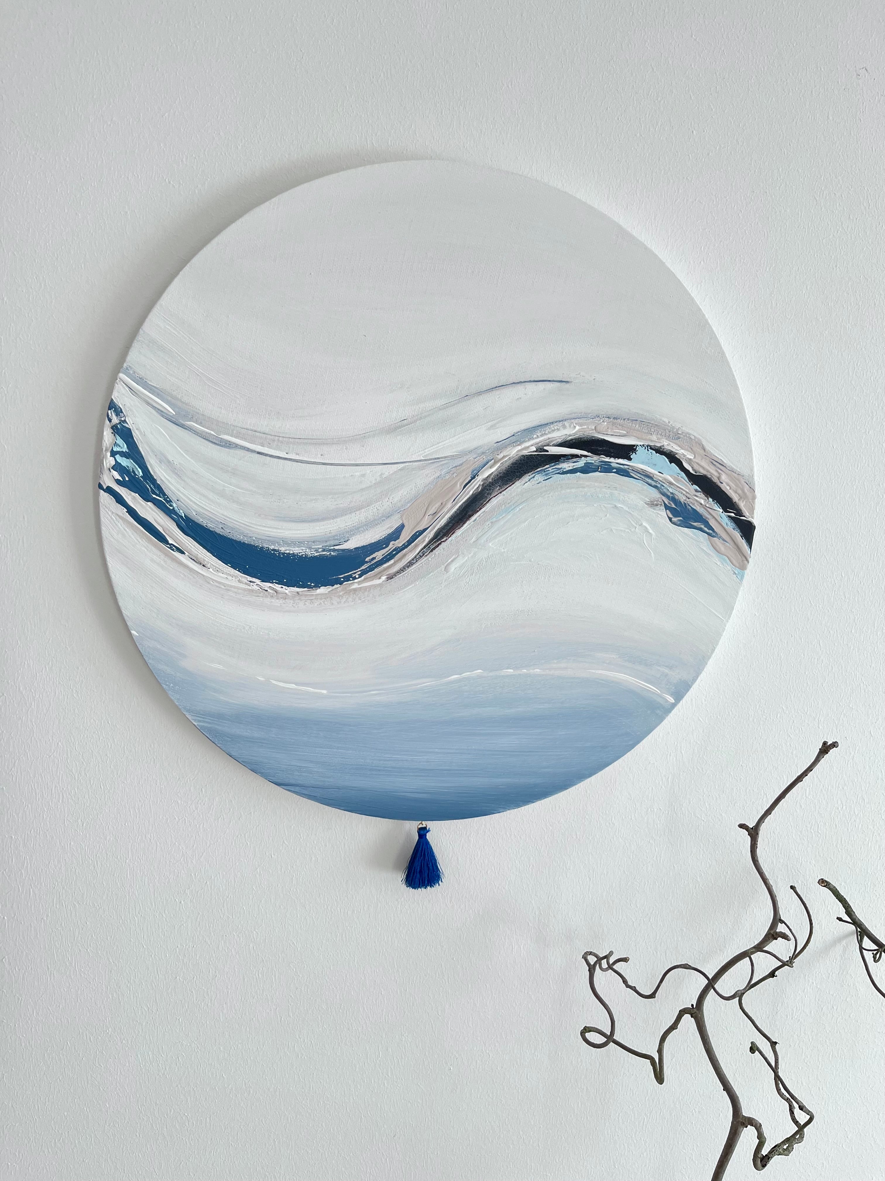 Das Kunstwerk "Cold Clarity". Die runde Leinwand ist oben sehr hell in weiß gehalten und unten in einem sanften gedeckten blau. Den sanften Abschluss bildet eine kleine blaue Quaste. Ein frostiger kühler Atemzug ist belebend und kann so klärend sein. Der belebende Atemzug ist in einem großen Schwung durch weiches Kunstleder rechts und blauen und nude Tönen in dickerem Farbauftrag links auf dem Werk entstanden.