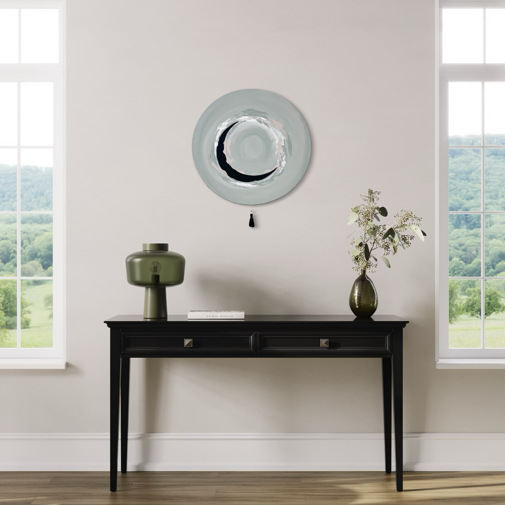 Das Kunstwerk "natural circle". Moderne Wabi Sabi Kunst mit daoistischer Seele von Nadine Elsner. 60cm im Durchmesser. Weiche graugrün, weiß und nude Töne mit weichem Kunstleder im Halbkreis. Sanfter Abschluss mit einer schwarzen  Quaste. Das Bild ergänzt ein einen hellen Raum mit großen Fenstern links und rechts neben dem Bild. Darunter steht ein schwarzer schöner Tisch. Der Ausblick geht in die Weite, alles sehr grün. Das Bild öffnet einen natürlichen Raum. Es bringt die Natur von außen nach innen.