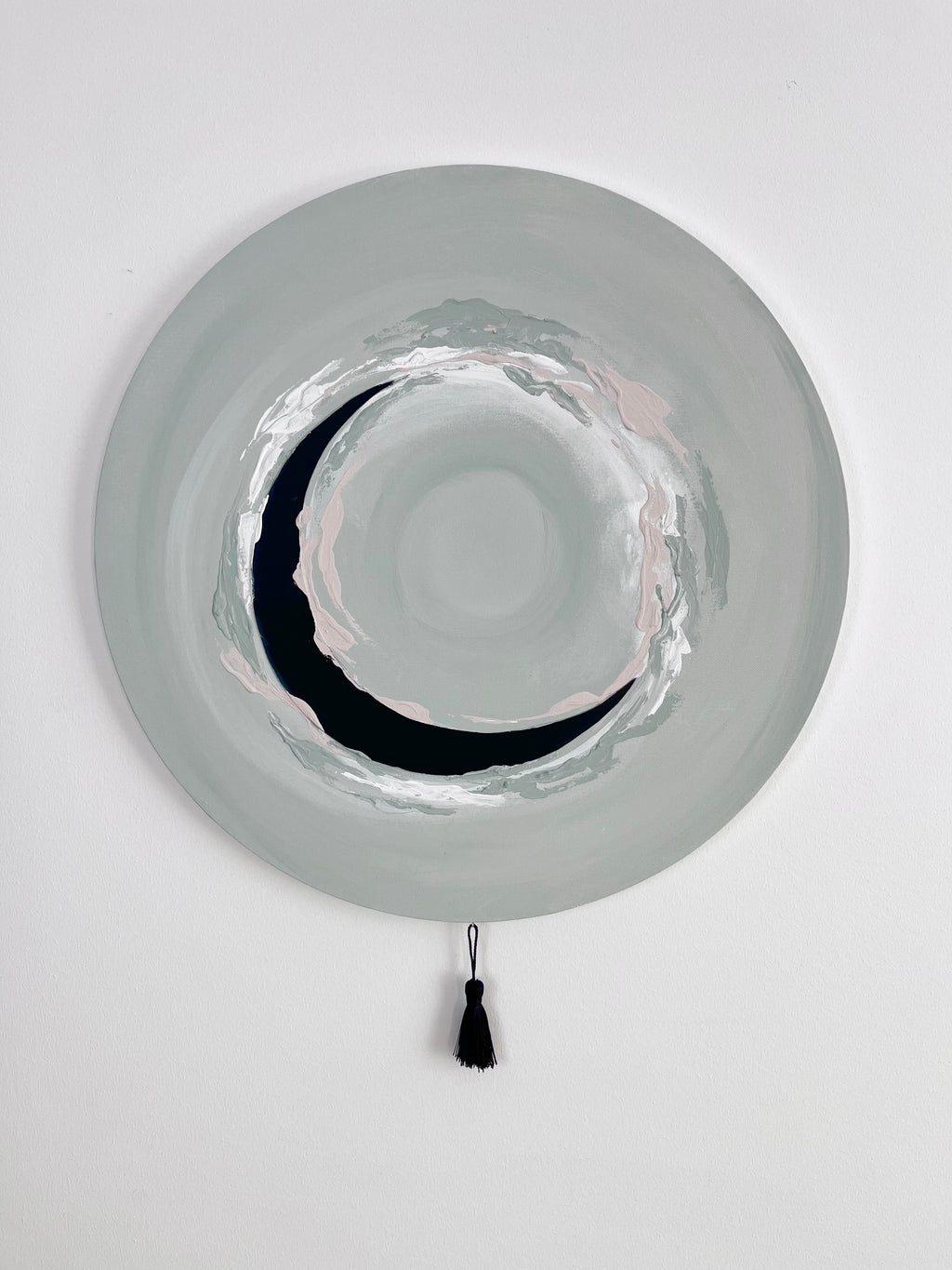 Das Kunstwerk "Natural Circle". Ein Bild in zartem grau grün im Hintergrund. In leichten sanften Verläufen ist ein Kreis angedeutet. Zur Mitte hin wird der Kreis durch einen schwarzen Halbkreis, wie eine Mondsichel aus Kunstleder ersichtlich. Der Zyklus schließt sich leicht durch dickere weiße, grau grüne und nude Farbene Farbaufträge durch Pinsel uns Spachtel. Den sanften Abschluss des runden Werkes, bildet eine größere schwarze Quaste.