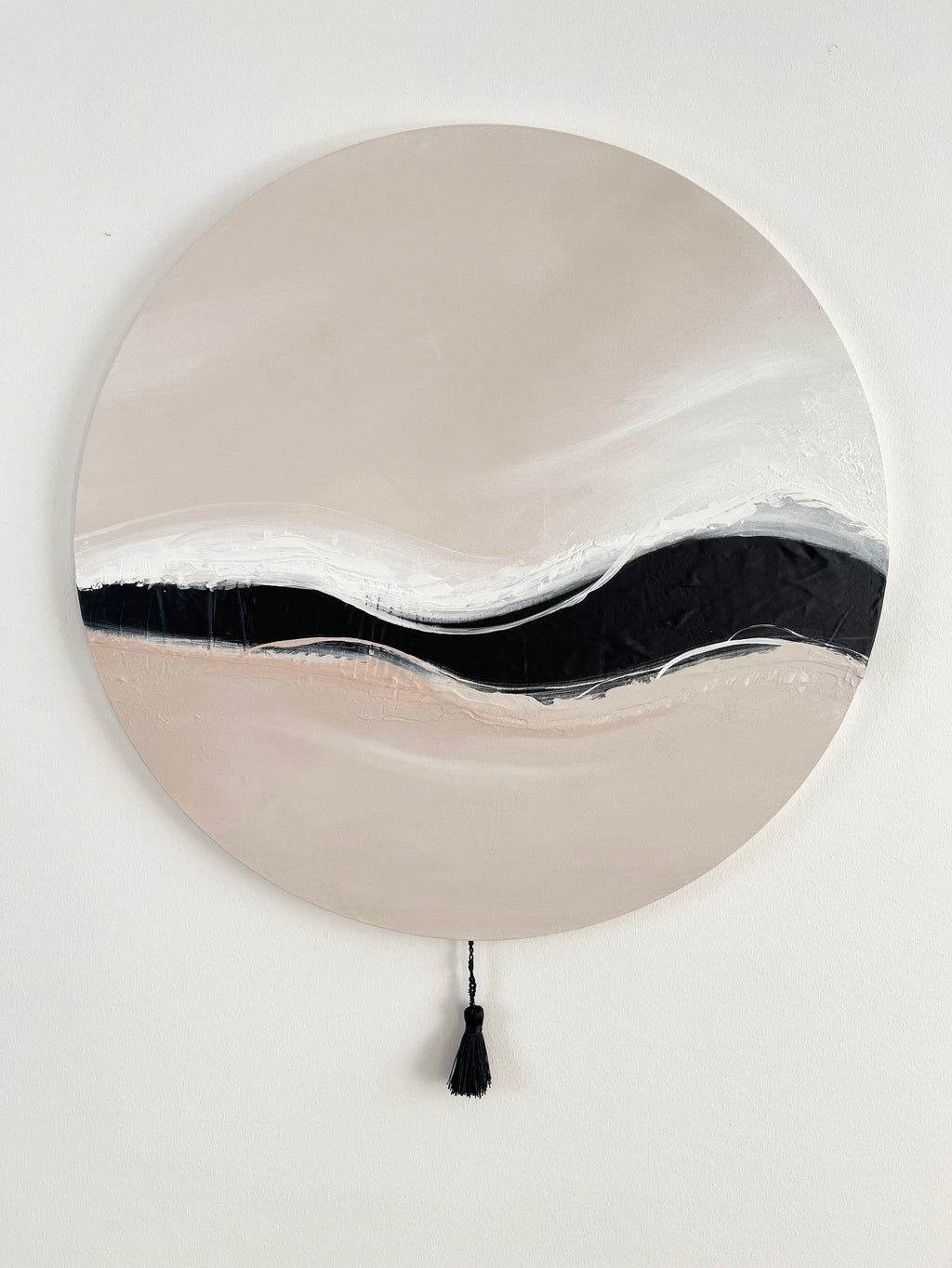 Das Kunstwerk "Warm Wave". Die gesamte Leinwand ist in zartem nude Ton und sanften Übergängen ins weiß gehalten. Durch die Mitte der runden Leinwand, zieht sich ein breiterer schwarzer Kunstlederstreifen. Oben und unten wird er durch zarte Pinselstriche weißer Wellen angeschnitten. Den sanften Abschluss bildet eine größere schwarze Quaste.