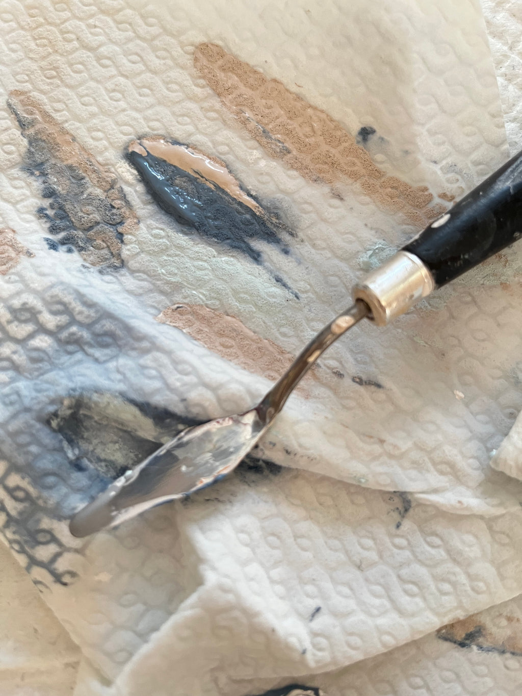Detailaufnahme des Materials im Atelier. Hier ein Tuch auf dem ein kleiner Spachtel liegt. Auf dem Tuch sind abgestrichene Farbspuren in grau blau und nude Tönen.